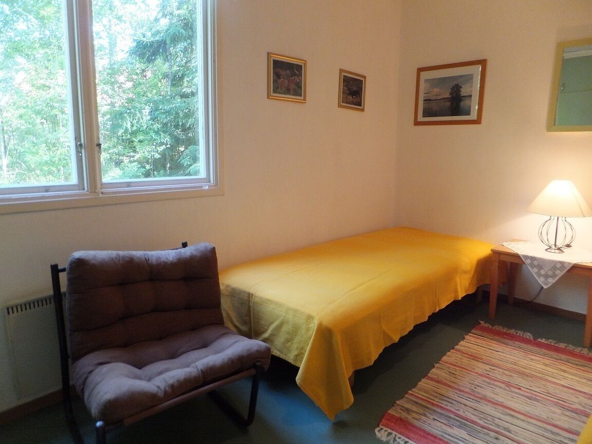 Ein kleines Zimmer mit einem Bett, einem Sessel, einem Tisch mit einer Lampe und zwei gerahmten Bildern an der Wand.