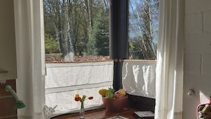 A kitchen with a window view of a forest, a sink, a small table with a vase of flowers, and a dish rack. - Apartment Berg & Barkenhoff vis-à-vis the Barkenhoff for 1-5 people (Worpswede)