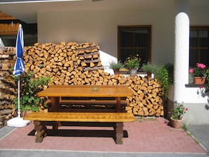 A wooden picnic table with benches, a stack of firewood, and potted plants against a building. - Mill - Blue Silvretta (Galtür)