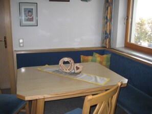 A room with a wooden table, blue cushioned bench, and a window with curtains. - Mill - Blue Silvretta (Galtür)