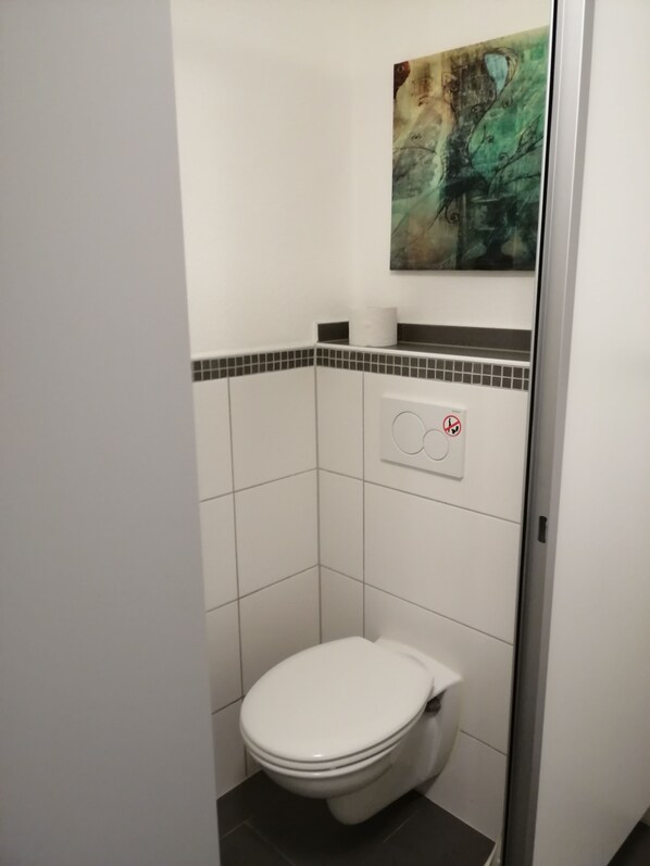 A small, white bathroom with a toilet, a wall-mounted paper towel dispenser, and a painting above the toilet. - Guest / fitter room ADES HAUS - uncomfortable somewhere else! (Überherrn)