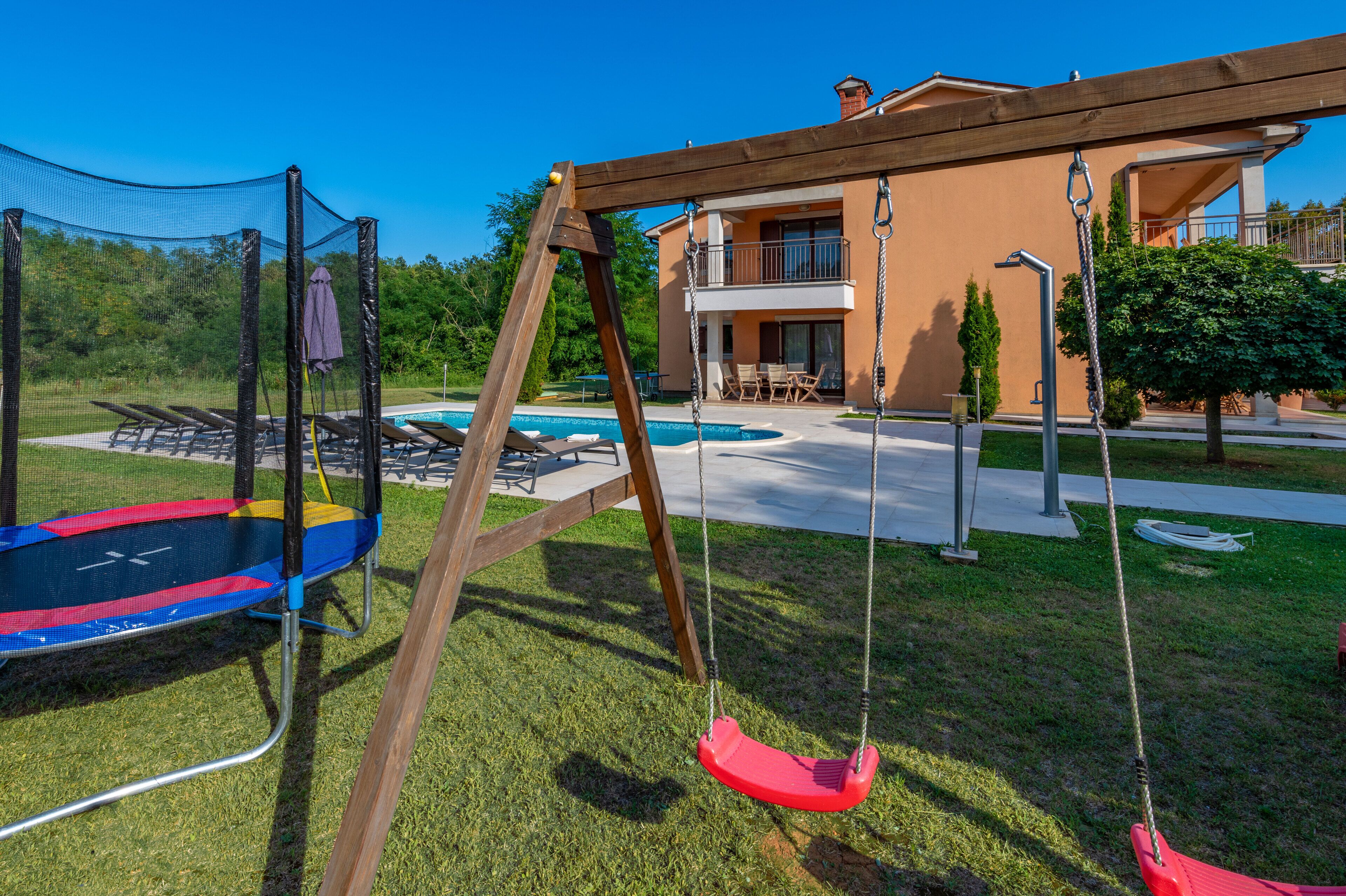 A trampoline and swing set in a backyard with a pool and a two-story building in the background.