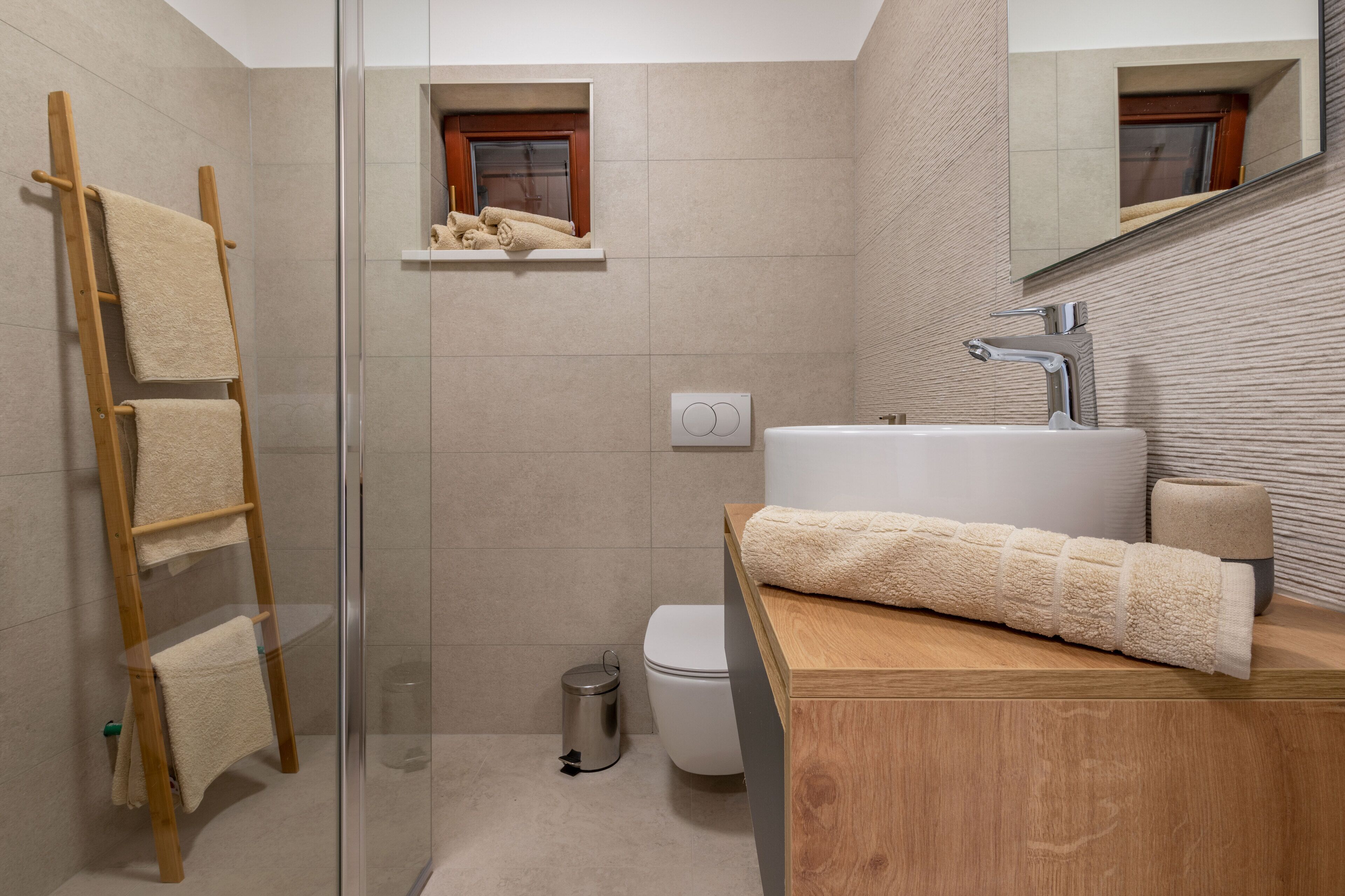 Een moderne badkamer met een douchecabine van glas, een houten handdoekrek, een witte wasbak en een klein raam boven het toilet.
