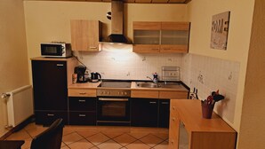 Cozinha compacta com micro-ondas, máquina de café e fogão. Há uma pia com torneira e um painel de azulejos. A cozinha possui armários de madeira e piso de azulejo.
