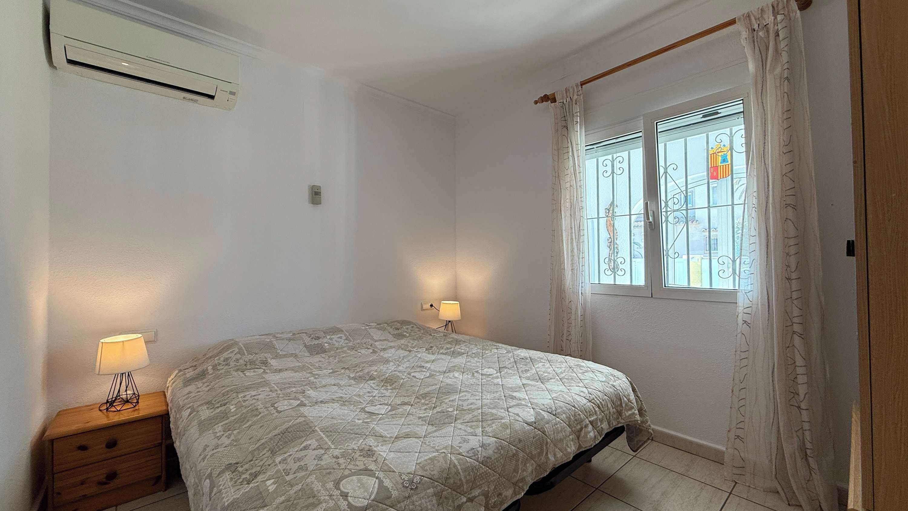 Et soveværelse med seng, sengeborde, et vindue med dekorativt jerngitter og en aircondition.