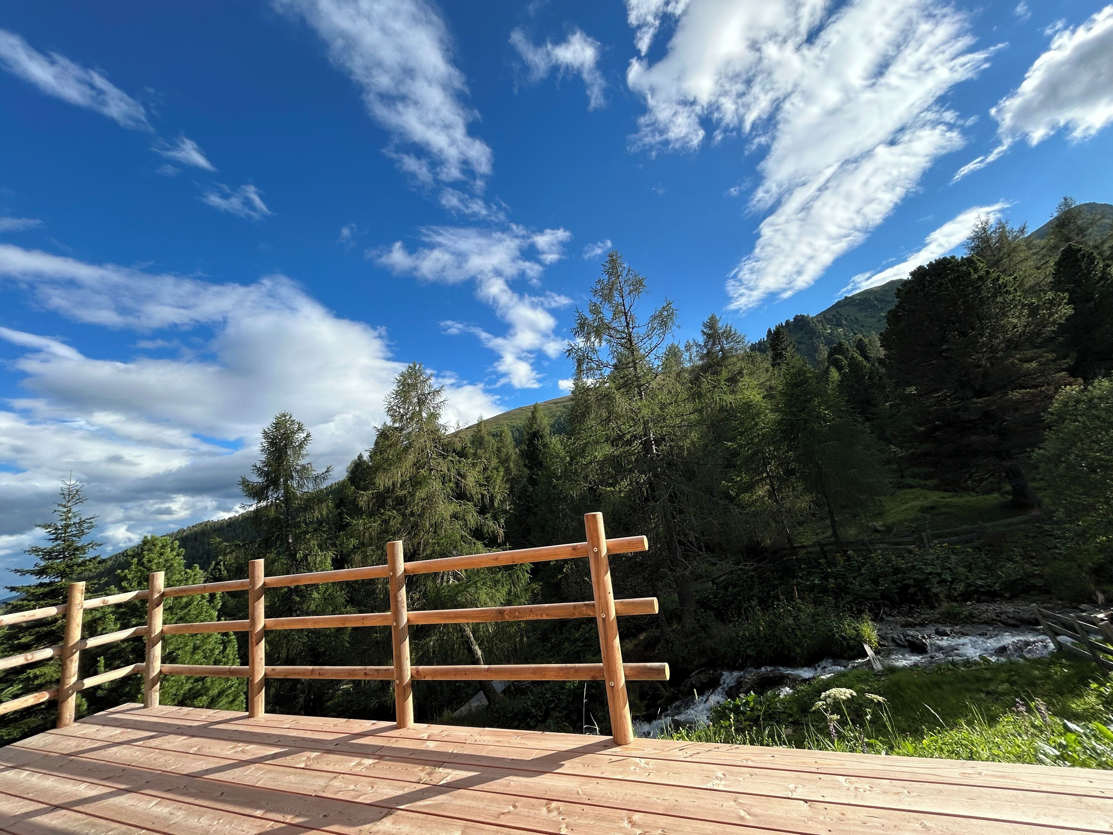 A wooden deck with a railing overlooking a forested area and a stream.