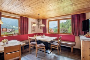 A dining area with a wooden table, chairs, and a bench. There are windows with curtains, a wall-mounted lamp, and a television.