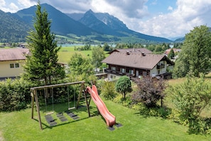 Zona residencial con un parque infantil que incluye un tobogán, rodeada de abundante vegetación y montañas al fondo.