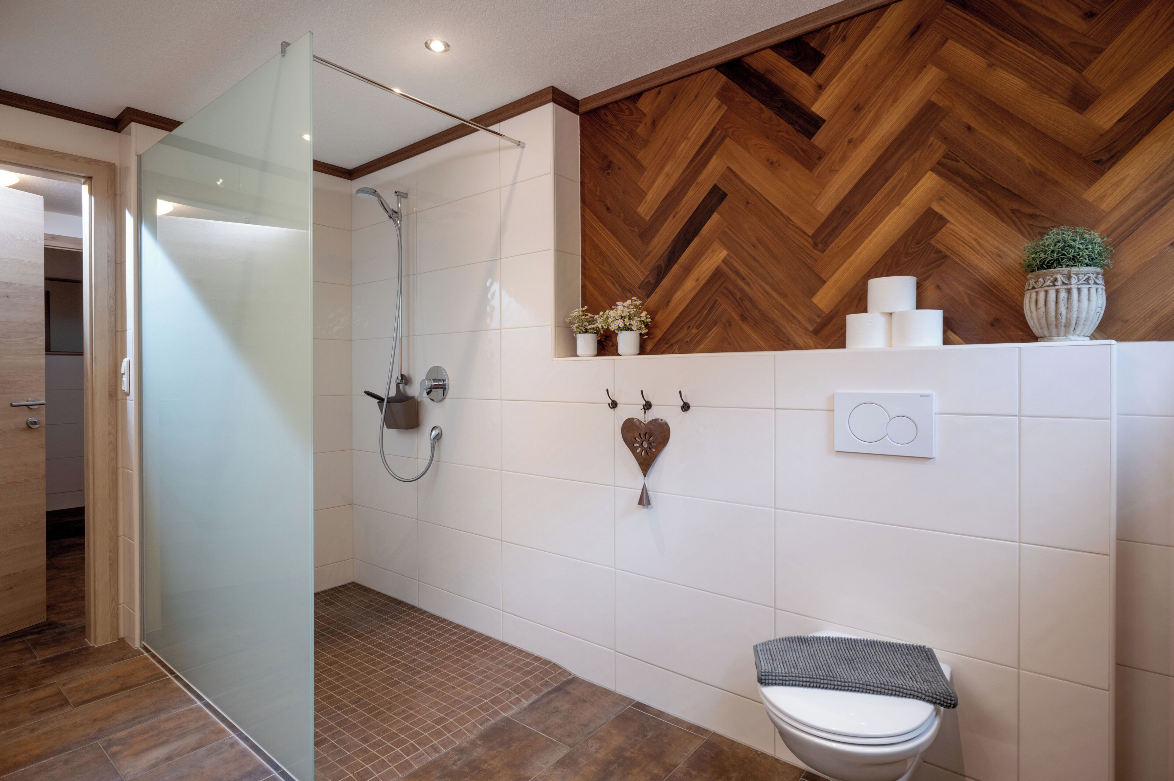 A modern bathroom with a glass shower enclosure, a wooden herringbone wall, a toilet, and a small potted plant on the wall.