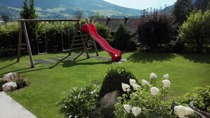A playground with a red slide and swings, surrounded by green grass and flowering plants, with mountains in the background.
