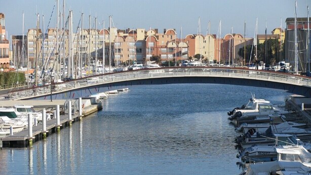 A marina with boats docked, a curved bridge, and buildings in the background. - Exclusively furnished apartment, 3 minutes from the beach (Gruissan)