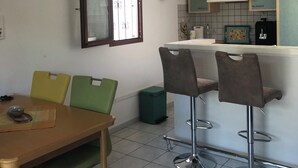 A compact kitchen with a dining area. It includes a table with chairs, a bar stool, and a small trash bin. A clock is mounted on the wall above the kitchen counter. - Exclusively furnished apartment, 3 minutes from the beach (Gruissan)