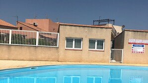 A swimming pool with a clear blue water surface, surrounded by a white fence. In the background, there is a building with a sign that reads "PISCINE" and another sign with "DOGS" and "NO DOGS" instructions. - Exclusively furnished apartment, 3 minutes from the beach (Gruissan)