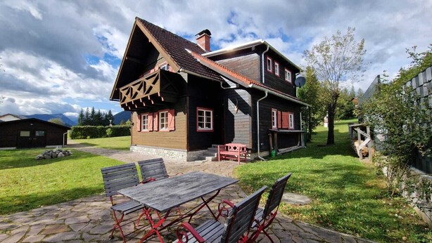 A traditional wooden house with a sloped roof, a stone patio with metal chairs, and a stone wall in the background. - cozy house in a sunny place with a fantastic mountain panorama (Bad Mitterndorf)