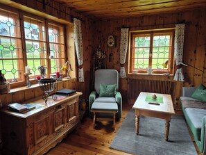 A cozy cabin interior with wooden walls, a wooden dining table, a cushioned armchair, a sofa, and a window with a view of greenery.
