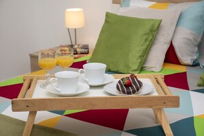 A breakfast tray with two glasses of orange juice, two white cups, and a chocolate-covered donut with a strawberry on a colorful geometric-patterned bedspread.