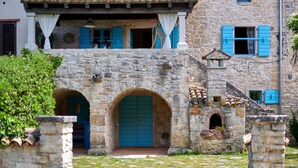 A stone building with a blue door and arched entrance, featuring a balcony with blue shutters and a tiled roof.
