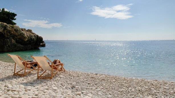 A beach with two wooden lounge chairs facing the sea, surrounded by pebbles.
