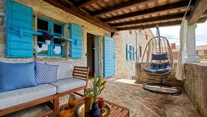 A stone building with blue shutters, a wooden sofa, a hanging chair, and a table with a tray of drinks and flowers.