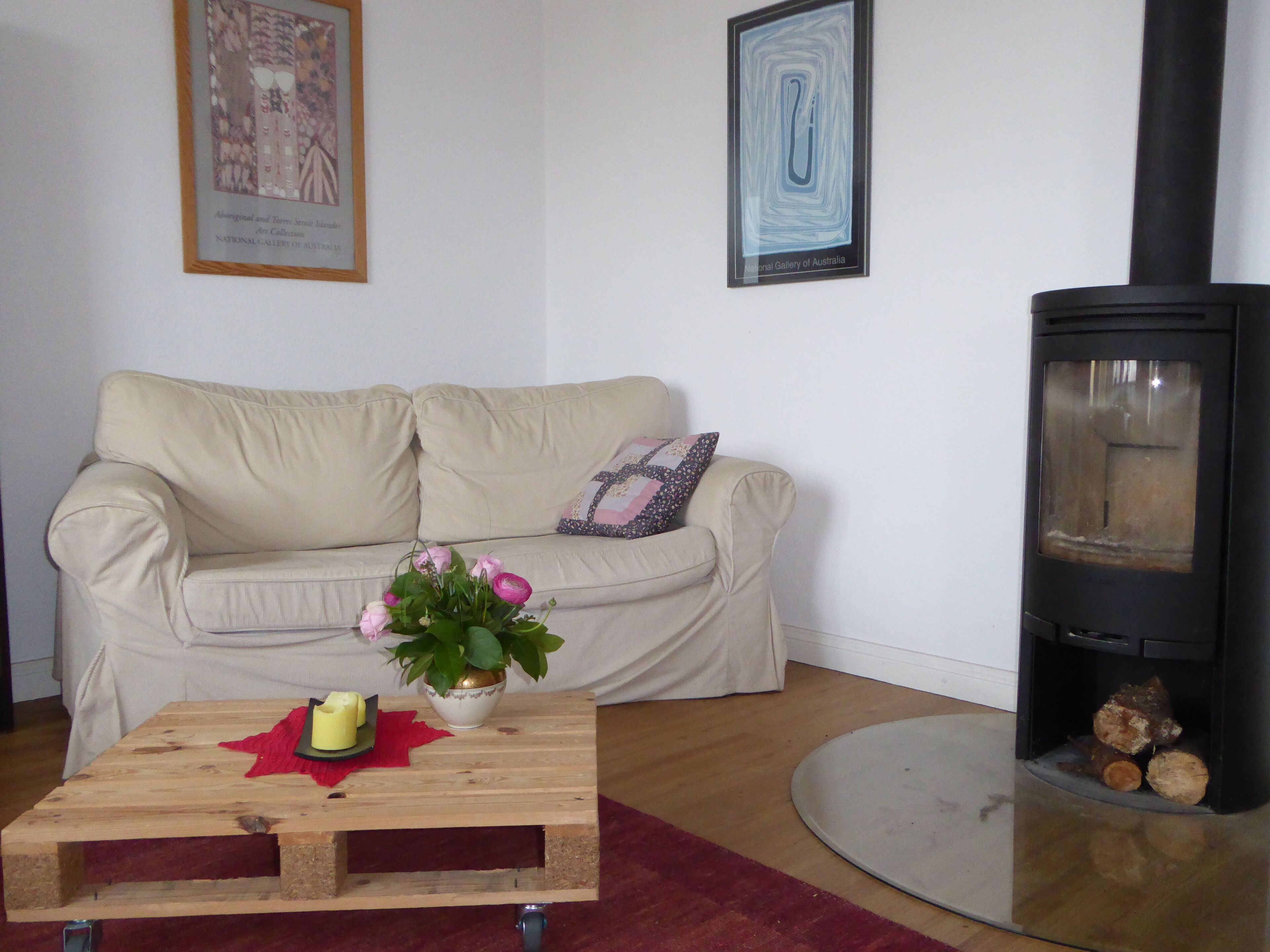 A living room with a beige sofa, a wooden coffee table with a vase of flowers, a framed picture on the wall, and a wood-burning stove.
