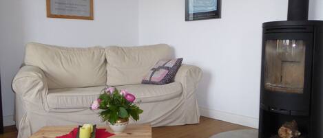 A living room with a beige sofa, a wooden coffee table with a vase of flowers, a framed picture on the wall, and a wood-burning stove.