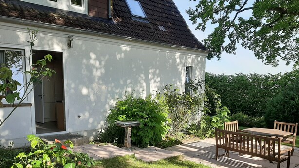 A white house with a tiled roof, a patio with a table and chairs, and a garden with various plants. - Enjoy stunning views over Kiel and nature (Kiel)