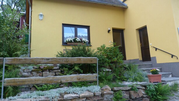 A yellow building with a stone wall, wooden steps, and a window with flowers. - Comfortable summer cottage in a beautiful surrounding (Marienberg)