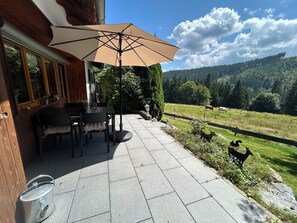 An outdoor patio with a table and chairs, a large umbrella, and a scenic view of a forested hillside.