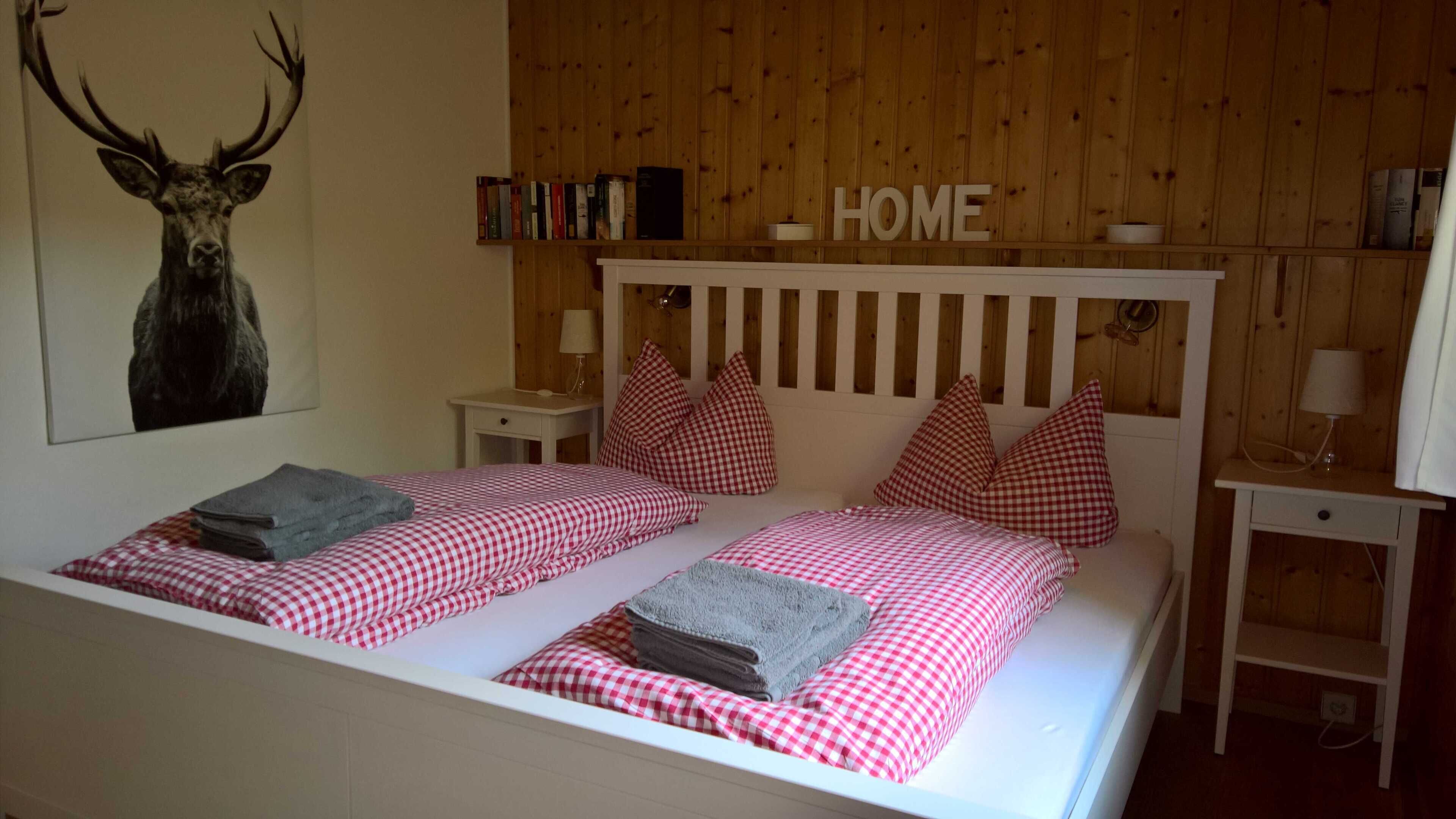 A bedroom with a wooden headboard, two beds with checkered bedding, pillows, a bedside table with a lamp, and a bookshelf.