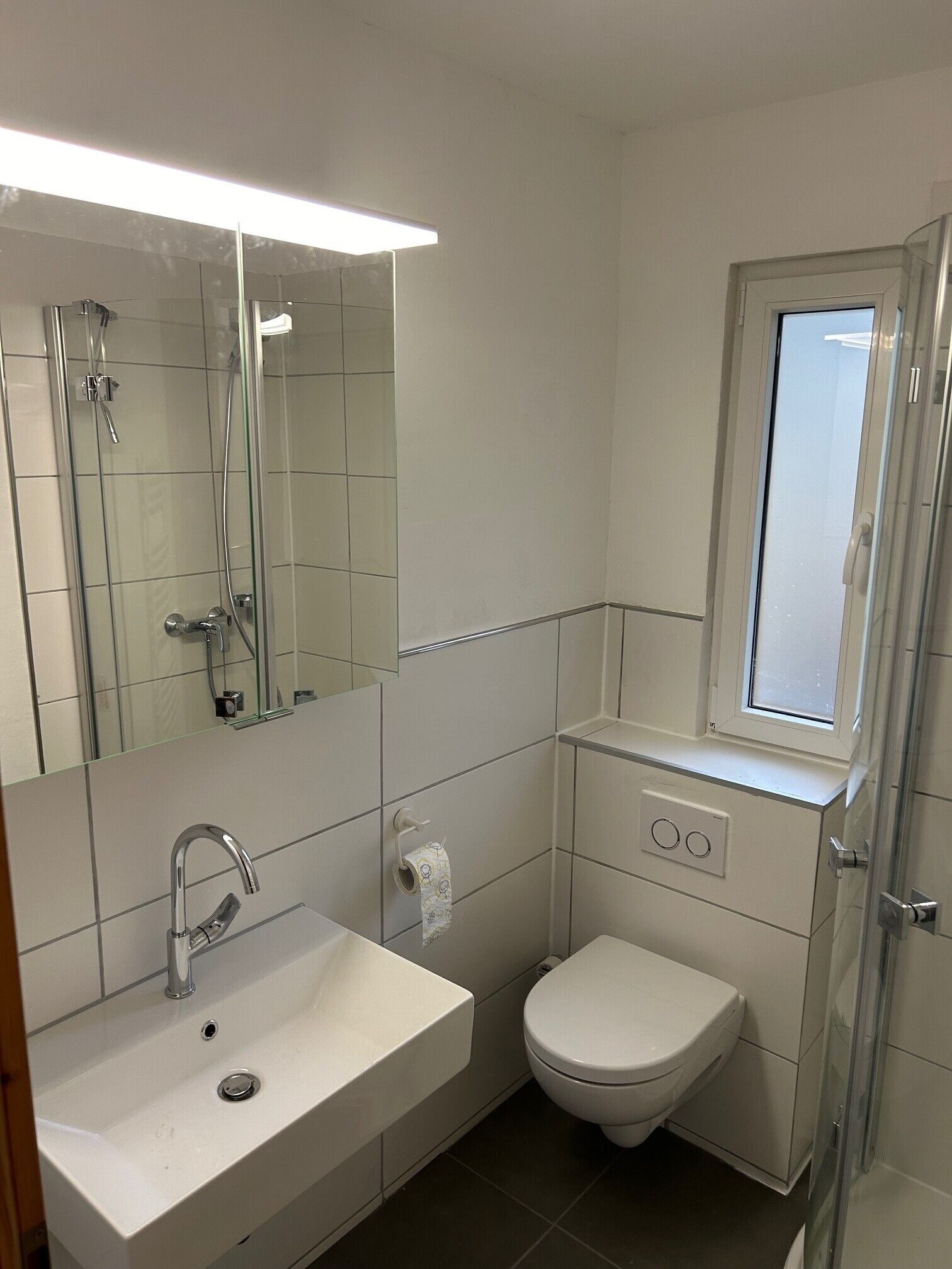 A modern bathroom with a white sink, a toilet, a glass shower enclosure, and a window.