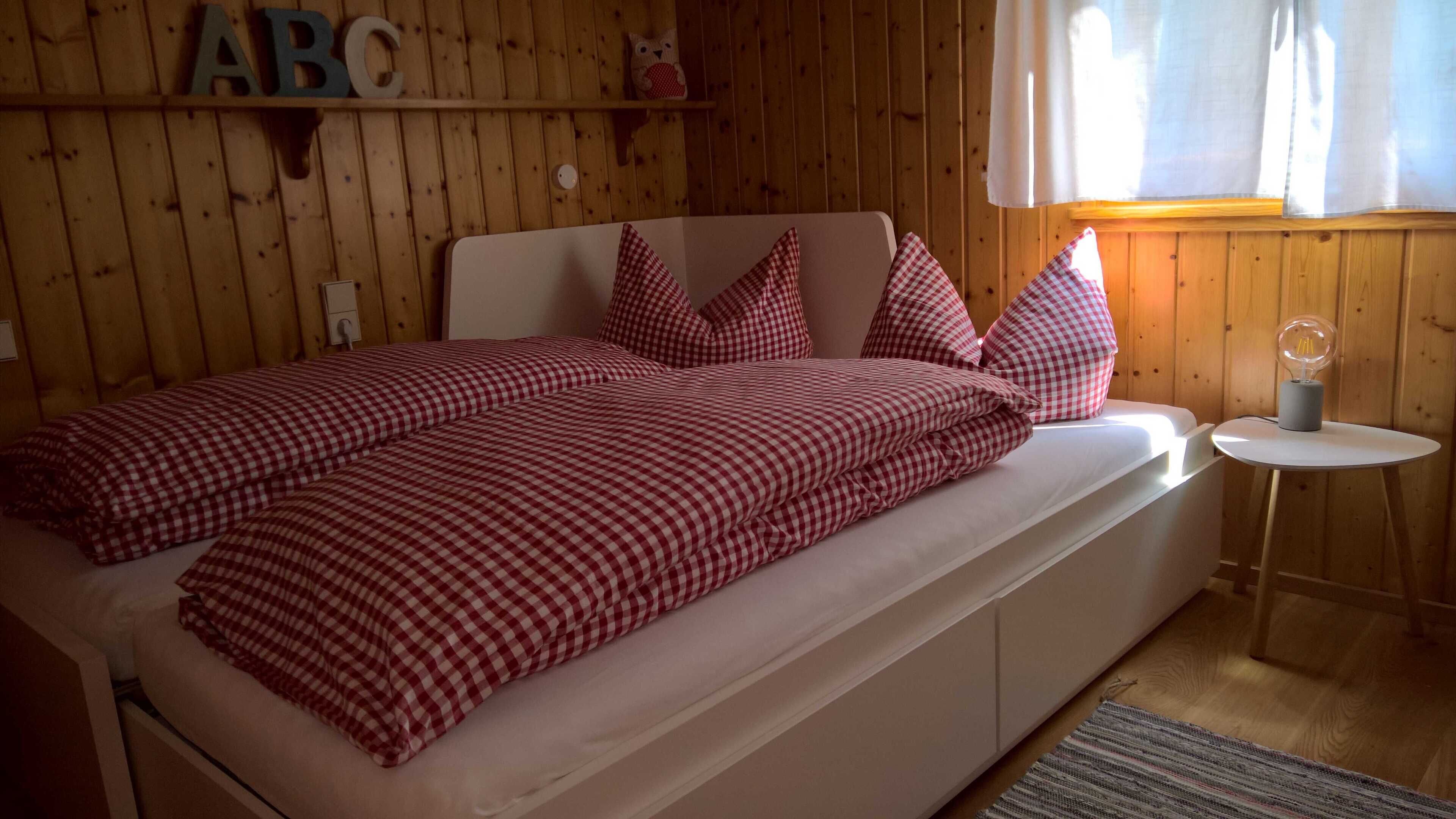 Sängyssä on ruutukuviollinen peitto ja tyynyjä, pieni pyöreä pöytä, jossa on lamppu, ja seinät ovat puuta, ja sängyn yläpuolella on hylly.