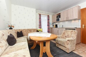 A compact living space with a sofa, a small round table, a kitchen area with cabinets and appliances, and a window with curtains.