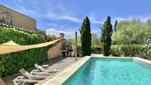 Une piscine rectangulaire à l’eau d’un bleu limpide, entourée d’une terrasse en pierre. On y trouve des chaises longues et une petite table sur laquelle est posé un chapeau.