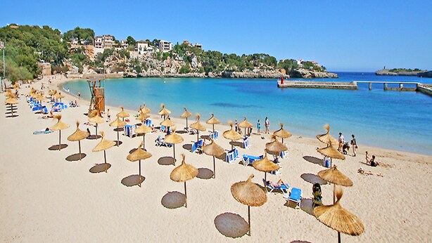 Une plage avec de nombreux parasols et des chaises longues, une mer d’un bleu limpide et des bâtiments en arrière-plan.