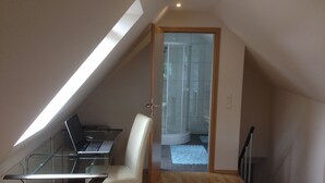 A small, modern room with a sloped ceiling, a glass desk with a laptop, a white chair, and a shower area visible through a glass door.