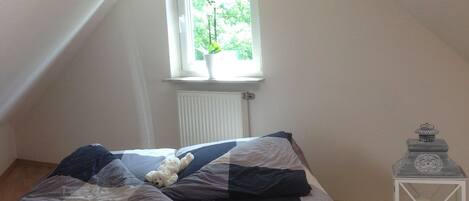 A bedroom with a sloped ceiling, a window with a view of greenery, a bed with a striped blanket, a white teddy bear, and a white lantern on a side table.
