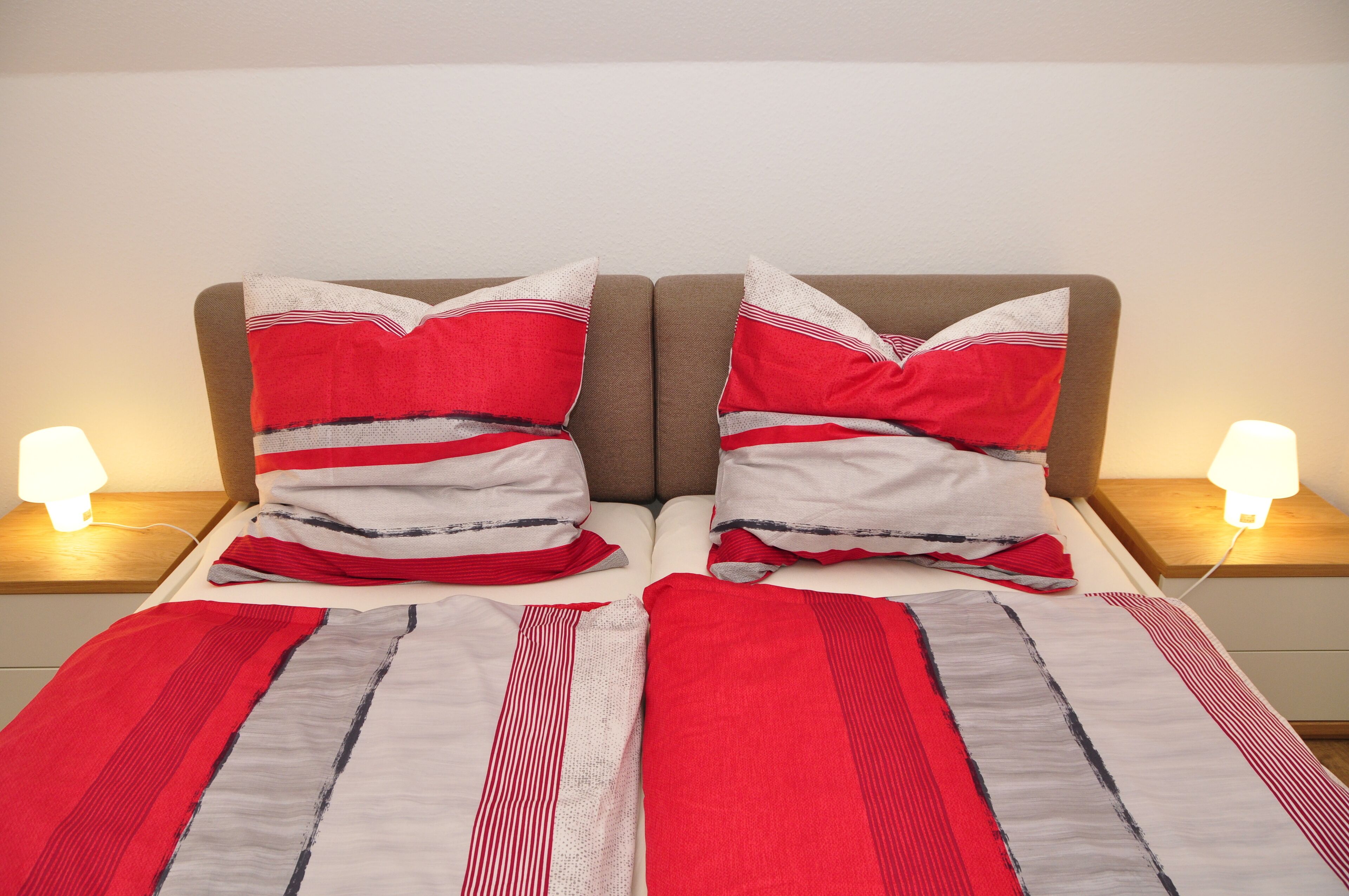 Een bed met rood-grijs gestreept beddengoed, twee kussens met rode en witte strepen, en bedlampen op houten nachtkastjes.
