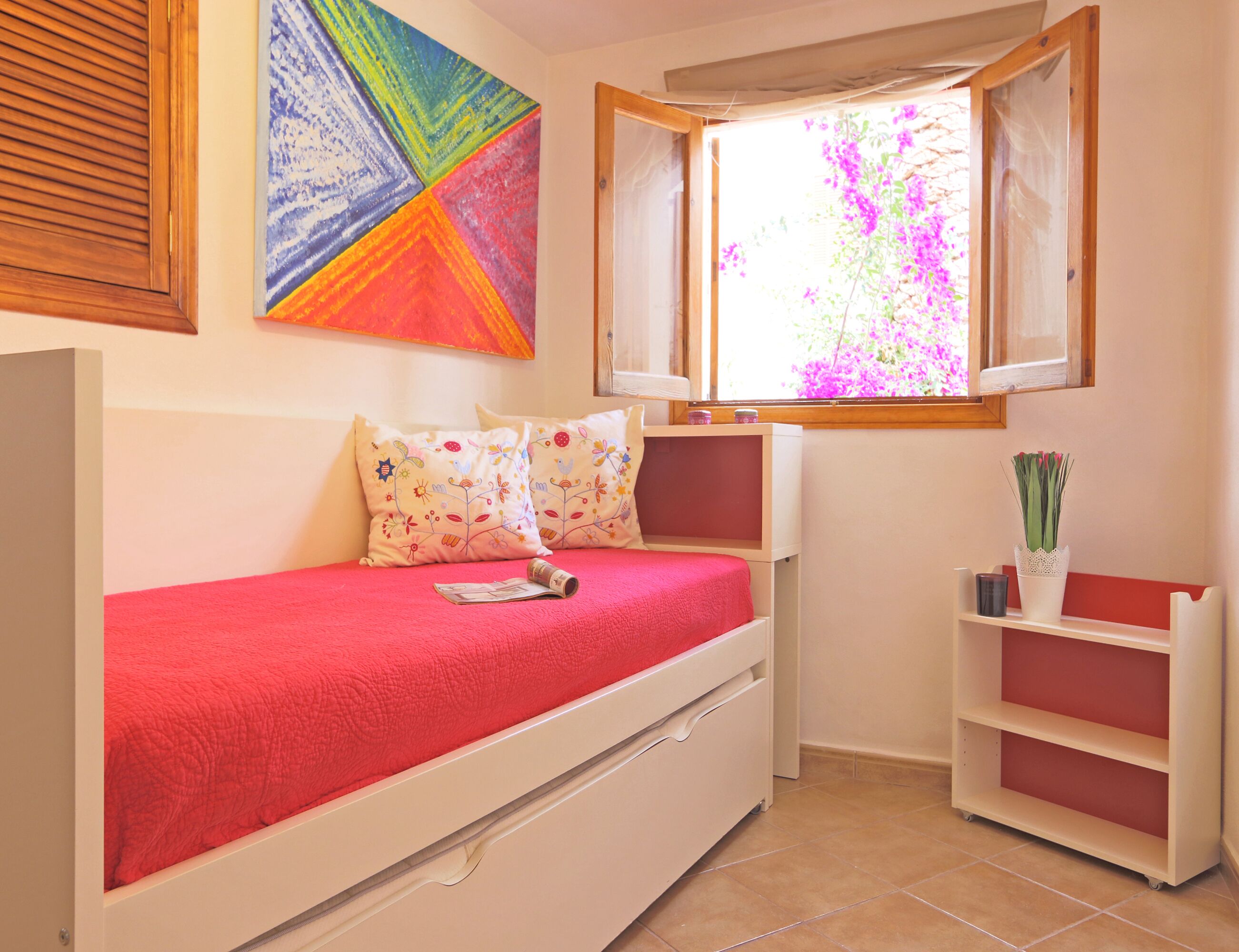 A small, cozy room with a single bed, a window with a view of purple flowers, a colorful abstract painting, and a small shelf with a potted plant.
