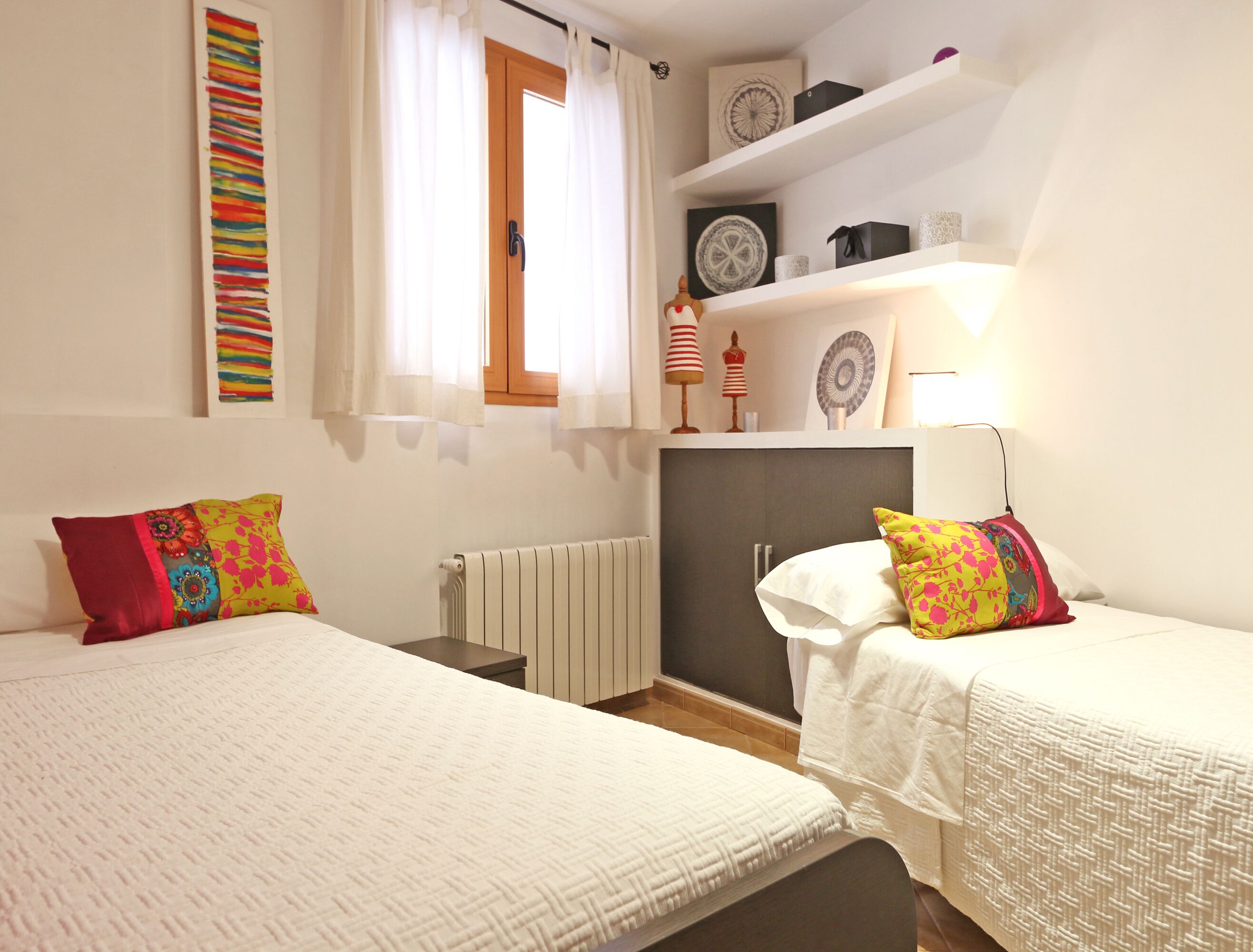 Un dormitori amb dos llits, un radiador, una estant amb objectes decoratius i una finestra amb cortines.