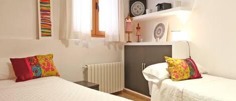 Un dormitori amb dos llits, un radiador, una estant amb objectes decoratius i una finestra amb cortines.