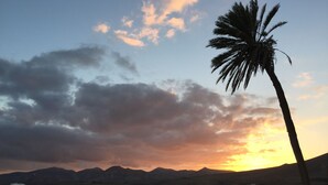 Une vue sur le coucher de soleil depuis un balcon, avec un palmier, des montagnes en arrière-plan et des bâtiments blancs.
