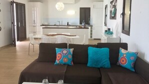 Un salon moderne avec un canapé marron, des coussins colorés, une table basse blanche et un coin repas comprenant une table et des chaises blanches.