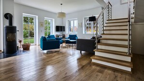 A spacious living room with a wooden floor, a black fireplace, blue sofas, and a staircase with a metal railing. - generous thatched cottage (160 m²) on the Baltic Sea, sauna, stove, forest edge location (Rerik)