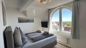 A modern bedroom with a large window, a ceiling fan, a flat-screen TV, and a bed with pillows and blankets. - WoogeDiva - the highest apartment on Wangerooge (Wangerooge)