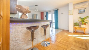 Une cuisine moderne avec un mur recouvert de pierres, deux tabourets de bar, un plancher en bois et une vue sur le salon, agrémenté d’une plante et d’un tableau.