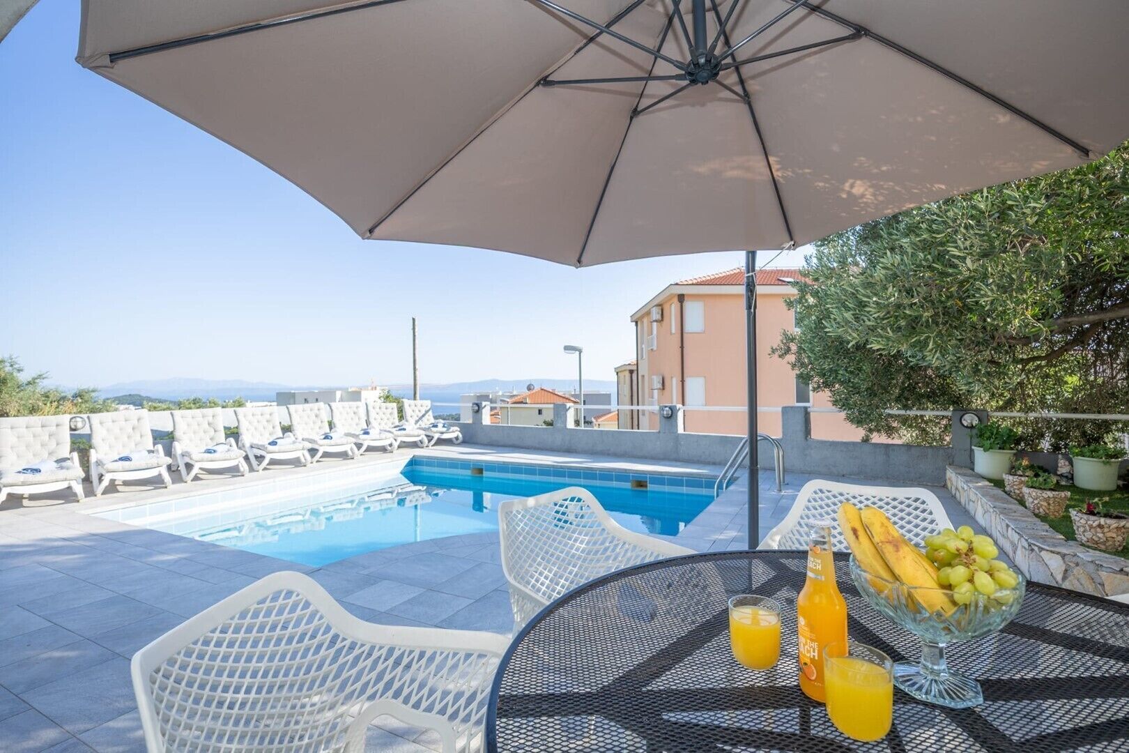 A poolside area with a view of the sea, lounge chairs, and a table with fruit and drinks.