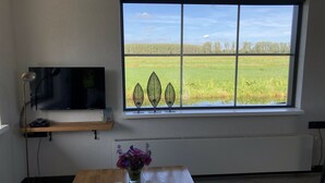 A room with a large window, a flat-screen TV on a shelf, a wooden table with a vase of flowers, and a patterned rug. - Nature house near Amsterdam Peace and beautiful view of private rental (Breukelen)