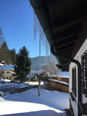 Paesaggio innevato con stalattiti che pendono da un tetto, una recinzione di legno e una casa con il tetto imbiancato dalla neve.