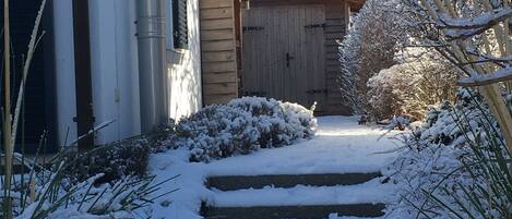 Un sentier enneigé mène à une porte en bois, bordé de plantes recouvertes de neige et d’un bâtiment dont on aperçoit les tuyaux et une fenêtre.