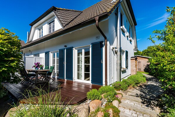 A two-story house with a wooden deck, a patio table with chairs, and a garden with rocks and plants.